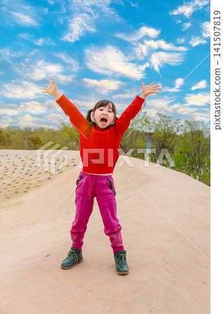 Happy Asian little girl jumping with arms raised outdoors under blue sky, joyful child playing in park 141507819