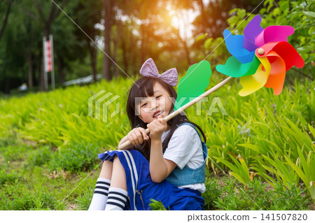 Happy Young East Asian Girl with Purple Bow Sitting in Sunny Green Meadow Holding Colorful Rainbow Pinwheel with Joyful Smile and Bright Summer Vibes. Happy Young East Asian Girl with Purple Bow Sitting in Sunny Green Meadow Holding Colorful Rainbow Pinwheel with Joyful Smile and Bright Summer Vibes. 141507820