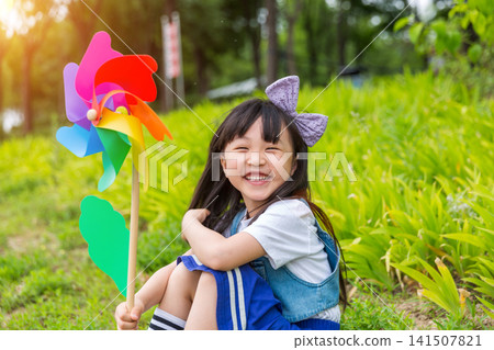 Happy Young East Asian Girl with Purple Bow Sitting in Sunny Green Meadow Holding Colorful Rainbow Pinwheel with Joyful Smile and Bright Summer Vibes. Happy Young East Asian Girl with Purple Bow Sitting in Sunny Green Meadow Holding Colorful Rainbow Pinwheel with Joyful Smile and Bright Summer Vibes. 141507821