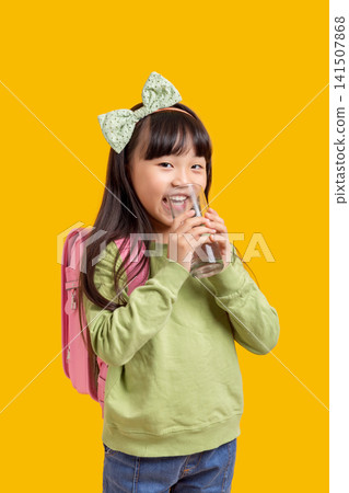 Cute Asian schoolgirl drinking water from glass, healthy hydration concept on yellow background 141507868