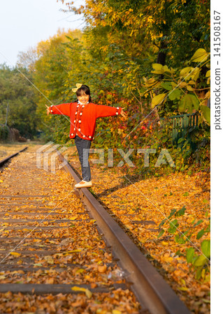 Young girl wearing a bright red sweater balances carefully on a single rail under warm autumn sunlight. Young girl wearing a bright red sweater balances carefully on a single rail under warm autumn sunlight. 141508167