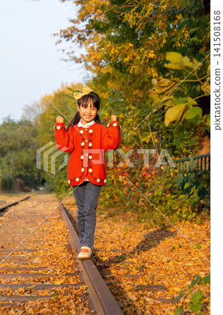 Young girl wearing a bright red sweater balances carefully on a single rail under warm autumn sunlight. Young girl wearing a bright red sweater balances carefully on a single rail under warm autumn sunlight. 141508168