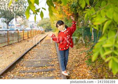 Young girl wearing a bright red sweater balances carefully on a single rail under warm autumn sunlight. Young girl wearing a bright red sweater balances carefully on a single rail under warm autumn sunlight. 141508169