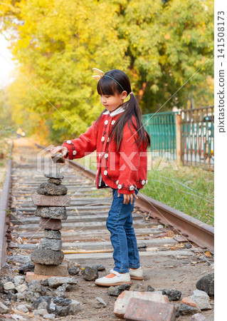 Curious little girl looks at a stack of balanced stones next to an old railway track in nature. Curious little girl looks at a stack of balanced stones next to an old railway track in nature. 141508173