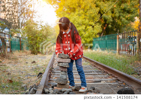Curious little girl looks at a stack of balanced stones next to an old railway track in nature. Curious little girl looks at a stack of balanced stones next to an old railway track in nature. 141508174