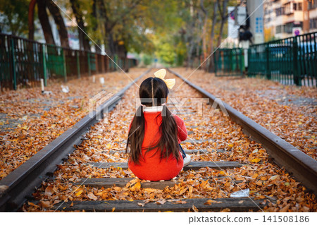 A little girl's silhouette sitting on the railway tracks, autumn leaves covering the ground. A little girl's silhouette sitting on the railway tracks, autumn leaves covering the ground. 141508186
