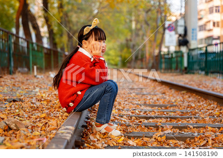 Little girl wearing a red sweater crouches on an old railway track ,On an autumn day Little girl wearing a red sweater crouches on an old railway track ,On an autumn day 141508190