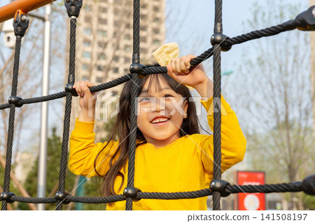 Joyful little girl smiles excitedly while playing inside a rope climbing net at an outdoor playground. Joyful little girl smiles excitedly while playing inside a rope climbing net at an outdoor playground. 141508197