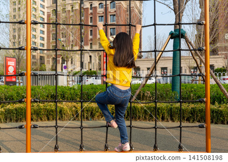 Full-length rear view of a young girl climbing up a large cargo rope net at an outdoor playground. Full-length rear view of a young girl climbing up a large cargo rope net at an outdoor playground. 141508198