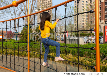 Full-length rear view of a young girl climbing up a large cargo rope net at an outdoor playground. Full-length rear view of a young girl climbing up a large cargo rope net at an outdoor playground. 141508199