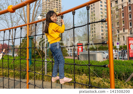 Full-length rear view of a young girl climbing up a large cargo rope net at an outdoor playground. 141508200