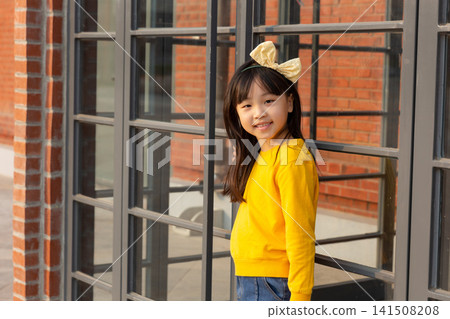 Cute young girl poses next to a large black-framed glass window with a brick wall background. Cute young girl poses next to a large black-framed glass window with a brick wall background. 141508208