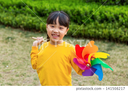 Medium shot of a bright-eyed young girl smiling warmly at the camera while holding a large rainbow pinwheel outdoors. Medium shot of a bright-eyed young girl smiling warmly at the camera while holding a large rainbow pinwheel outdoors. 141508217