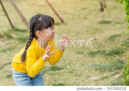 A playful little girl bends forward on a grassy lawn with a funny facial expression, trying to catch a flying butterfly. A playful little girl bends forward on a grassy lawn with a funny facial expression, trying to catch a flying butterfly. 141508218