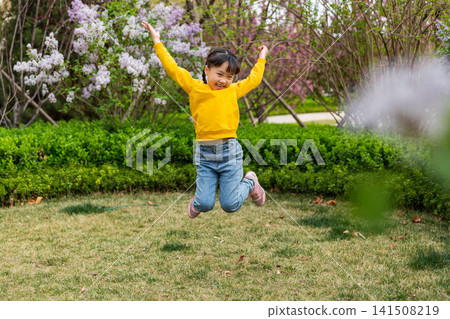 An energetic young girl mid-air jumps with arms raised happily on a park lawn with blooming white blossoms in the background. An energetic young girl mid-air jumps with arms raised happily on a park lawn with blooming white blossoms in the background. 141508219