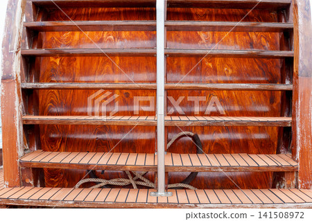 Close up of a polished wooden staircase on a luxury yacht featuring rich grain and teak decking steps. Nautical detail of ship architecture with a vertical metal handrail and rope below. Close up of a polished wooden staircase on a luxury yacht featuring rich grain and teak decking steps. Nautical detail of ship architecture with a vertical metal handrail and rope below. 141508972