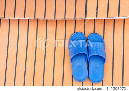 High angle view of blue rubber slippers on a wooden yacht deck with black caulking. This casual footwear represents summer relaxation, seaside vacation and nautical travel. Modern minimalist style. High angle view of blue rubber slippers on a wooden yacht deck with black caulking. This casual footwear represents summer relaxation, seaside vacation and nautical travel. Modern minimalist style. 141508973