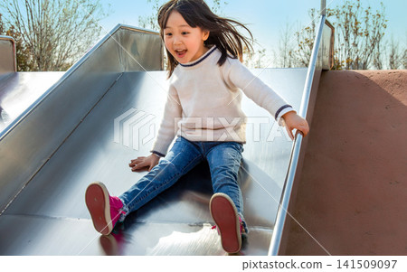 Excited little girl sliding down a metal slide at the playground on a sunny day Excited little girl sliding down a metal slide at the playground on a sunny day 141509097