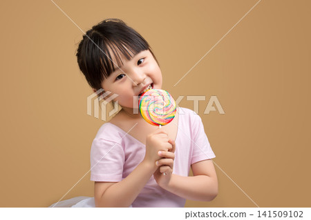 Asian Little Girl Enjoying Large Rainbow Lollipop on Beige Studio Background Asian Little Girl Enjoying Large Rainbow Lollipop on Beige Studio Background 141509102