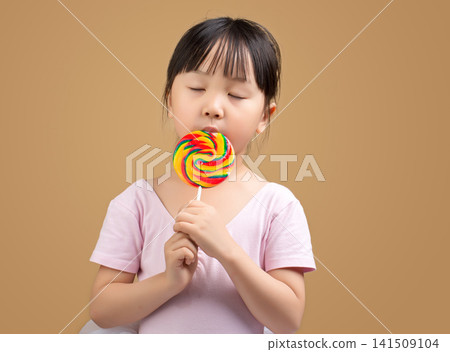 Asian Little Girl Eating Rainbow Lollipop with Eyes Closed on Beige Background 141509104