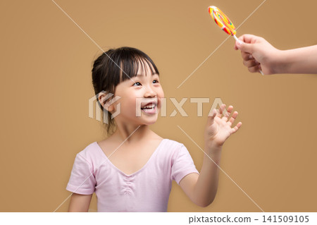 Happy Asian Little Girl Reaching for Colorful Lollipop Offered by Adult Hand on Beige Studio Background Happy Asian Little Girl Reaching for Colorful Lollipop Offered by Adult Hand on Beige Studio Background 141509105