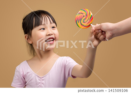Happy Asian Little Girl Reaching for Rainbow Lollipop Offered by Adult Hand Happy Asian Little Girl Reaching for Rainbow Lollipop Offered by Adult Hand 141509106