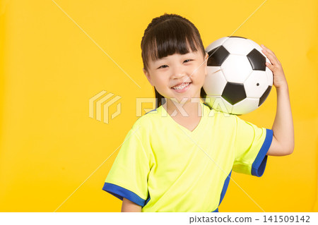 The smiling young girl leans slightly to the side on the yellow background, balancing the soccer ball casually behind her head with one hand, looking up playfully with a joyful expression in her eyes. 141509142