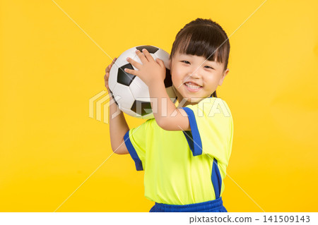 The smiling young girl leans slightly to the side on the yellow background, balancing the soccer ball casually behind her head with one hand, looking up playfully with a joyful expression in her eyes. The smiling young girl leans slightly to the side on the yellow background, balancing the soccer ball casually behind her head with one hand, looking up playfully with a joyful expression in her eyes. 141509143