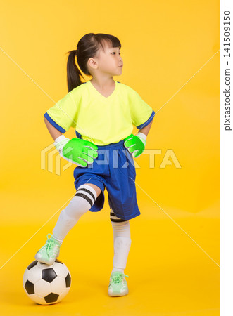 The young East Asian goalkeeper stands against the solid yellow wall, resting her right foot on the black and white ball. She wears green and white gloves, places both hands firmly on her hips The young East Asian goalkeeper stands against the solid yellow wall, resting her right foot on the black and white ball. She wears green and white gloves, places both hands firmly on her hips 141509150