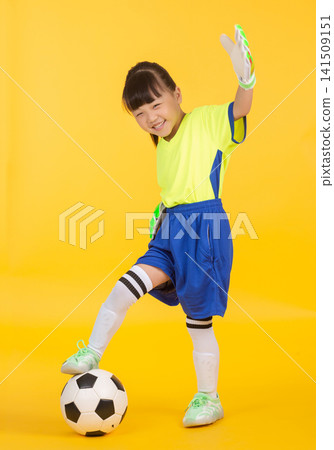a East Asian girl, now wearing bright green and white goalkeeper gloves, stands balanced with one foot on the soccer ball against the yellow studio wall. a East Asian girl, now wearing bright green and white goalkeeper gloves, stands balanced with one foot on the soccer ball against the yellow studio wall. 141509151