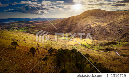 Sunlight streams over a mountain valley with a winding river running through grassy fields and scattered trees under a cloudy sky. 141510729