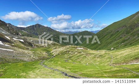 An alpine valley unfolds along the Roshka to Juta route in Georgia, where a narrow trail, melting snowfields, rocky slopes An alpine valley unfolds along the Roshka to Juta route in Georgia, where a narrow trail, melting snowfields, rocky slopes 141511094