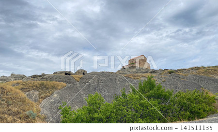 Uplistsikhe Church overlooks the rocky cave city in a wide low-angle view, framed by exposed stone, grass, green trees Uplistsikhe Church overlooks the rocky cave city in a wide low-angle view, framed by exposed stone, grass, green trees 141511135