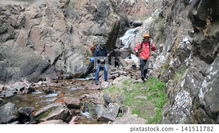 Two men are walking along a rocky path next to a stream. A hiking instructor accompanies a tourist on a hike. A trip to the mountains Two men are walking along a rocky path next to a stream. A hiking instructor accompanies a tourist on a hike. A trip to the mountains 141511782