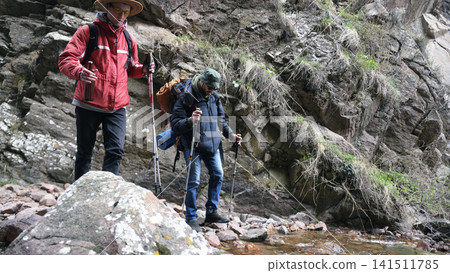 Tourists are walking in nature. Two guys are walking along a rocky path along a stream. A team of hikers crosses a small river Tourists are walking in nature. Two guys are walking along a rocky path along a stream. A team of hikers crosses a small river 141511785