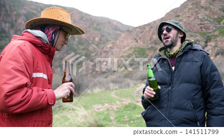 Two men are standing in a field surrounded by mountains and drinking beer. The guys are holding bottles of alcoholic beverage 141511799