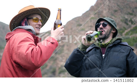 Two guys are getting drunk in the mountains. Friends drink beer in nature. Alcoholism as a bad habit. Rest after a hard day's work 141511802