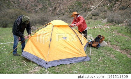 Two guys are attaching metal arcs to the tent. Men are setting up a bright orange tent in a grassy field. Trekking and traveling Two guys are attaching metal arcs to the tent. Men are setting up a bright orange tent in a grassy field. Trekking and traveling 141511809