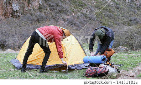 Two men set up a tent on a green lawn in the mountains. A summer hike. Tourists travel independently without a guide Two men set up a tent on a green lawn in the mountains. A summer hike. Tourists travel independently without a guide 141511818