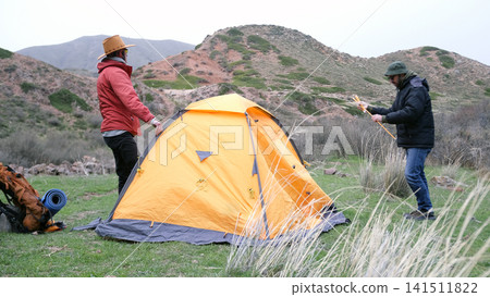 Life in a tent. Hiking in the mountains. Two men are standing next to an orange tent in a grassy field. A trip through the Asian mountains Life in a tent. Hiking in the mountains. Two men are standing next to an orange tent in a grassy field. A trip through the Asian mountains 141511822