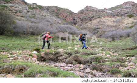 Two people are walking along a rocky path in a field among mountains and hills. A couple of friends are going on a little camping Two people are walking along a rocky path in a field among mountains and hills. A couple of friends are going on a little camping 141511853