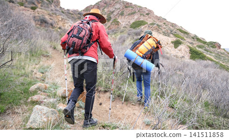 Two travelers climb the mountain with backpacks and trekking poles. The guide is taking his friend camping all weekend Two travelers climb the mountain with backpacks and trekking poles. The guide is taking his friend camping all weekend 141511858