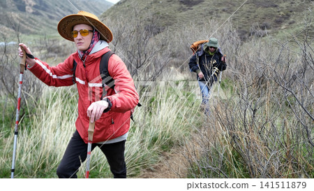 A man in a red jacket, yellow glasses and a cowboy hat walks along the trail with trekking poles. He is followed by his friend 141511879