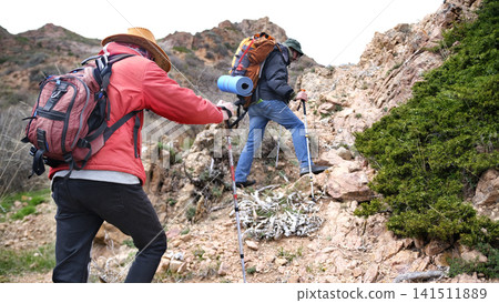 Tourists - climbers climb the mountain. Extreme and freedom. Two friends are walking up a mountain path past rocks and juniper bushes Tourists - climbers climb the mountain. Extreme and freedom. Two friends are walking up a mountain path past rocks and juniper bushes 141511889
