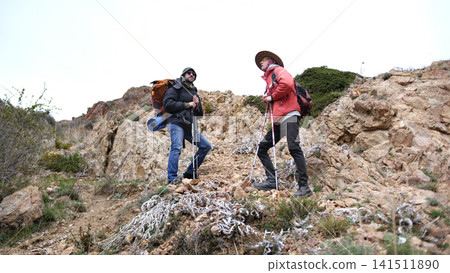 Two travelers are resting halfway up the mountain. The wanderers are standing on a rocky hill and recovering their breath after a hard climb Two travelers are resting halfway up the mountain. The wanderers are standing on a rocky hill and recovering their breath after a hard climb 141511890