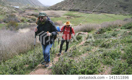 Tired travelers walk up the mountain path. Two men are traveling together. Trekking in the mountains. Tourists actively spend their weekends Tired travelers walk up the mountain path. Two men are traveling together. Trekking in the mountains. Tourists actively spend their weekends 141511896