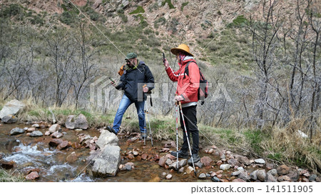 Two tourists are standing by the riverbed taking photos on their phones. The hikers went hiking in a mountain river 141511905