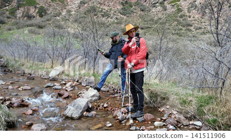 Two travelers stopped by a small mountain river to take a few photos on their phone. Two men are standing on a rocky riverbank Two travelers stopped by a small mountain river to take a few photos on their phone. Two men are standing on a rocky riverbank 141511906