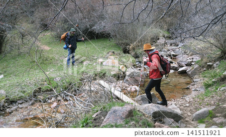 The guide watches his friend walk across a small river on a plank. Caring and mutual assistance. Trekking, hiking and hiking The guide watches his friend walk across a small river on a plank. Caring and mutual assistance. Trekking, hiking and hiking 141511921