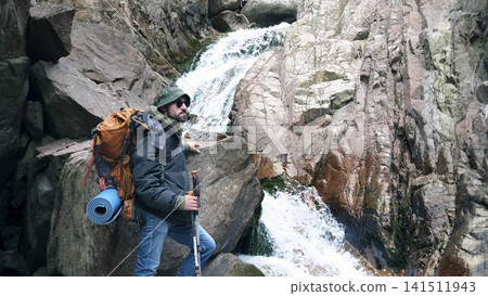 A lonely man in a panama hat, sunglasses, with a backpack and trekking poles stands in front of a waterfall and thinks about the eternal A lonely man in a panama hat, sunglasses, with a backpack and trekking poles stands in front of a waterfall and thinks about the eternal 141511943